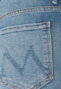 Primer plano de un bolsillo trasero de jeans de mezclilla azul con costuras naranja y un diseño bordado en forma de montaña.