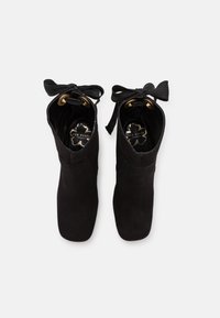 Pari saappaita mustasta mokasta, joissa on neliön muotoiset varpaat ja mustat nauhat, jotka on kuljetettu kullanväristen silmukoiden läpi, nähtynä ylhäältä.