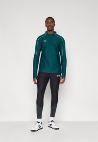 Τιρκουάζ μακρυμάνικη μπλούζα με φερμουάρ και πράσινες λεπτομέρειες, σε συνδυασμό με μαύρο στενό παντελόνι με υφασμένα κομμάτια στα κάτω μέρη και λευκά αθλητικά παπούτσια.