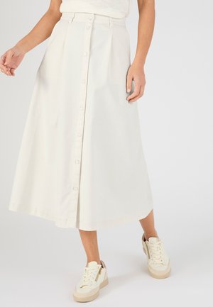 Frau trägt einen weißen, vorne geknöpften Midirock kombiniert mit weißen Sneakern und steht vor einem schlichten hellen Hintergrund.