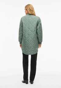 Žalių atsegamų siūlių striukė su stovu ir ilgomis rankovėmis, siekianti vidurį šlaunies. Dėvima virš juodų kelnių, atskleidžianti tekstūruotą dizainą.