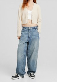 Cropped, light beige cardigan with button closure, paired with loose, light blue denim jeans. Black and white sneakers complete the outfit.