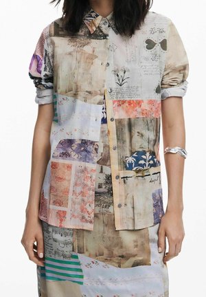 Person trägt ein geknöpftes, langärmeliges Hemd und einen Rock mit bunten, gemischten Patchworkmustern, die florale, schriftliche und abstrakte Designs zeigen.