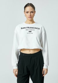 Witte cropped sweatshirt met de opdruk "SAN FRANCISCO" in het zwart, met lange mouwen en een ronde halslijn, in combinatie met zwarte high-waisted broek.