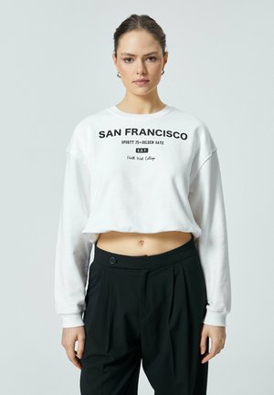 Valkoinen lyhyt svetari, jossa on mustalla painettu teksti "SAN FRANCISCO", pitkät hihat ja pyöreä kaula-aukko, yhdistettynä mustiin korkeavyötäröisiin housuihin.