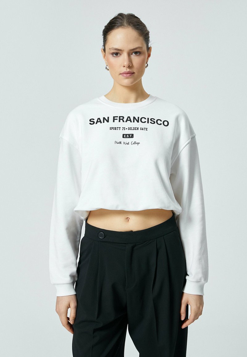 Witte cropped sweatshirt met de opdruk "SAN FRANCISCO" in het zwart, met lange mouwen en een ronde halslijn, in combinatie met zwarte high-waisted broek.
