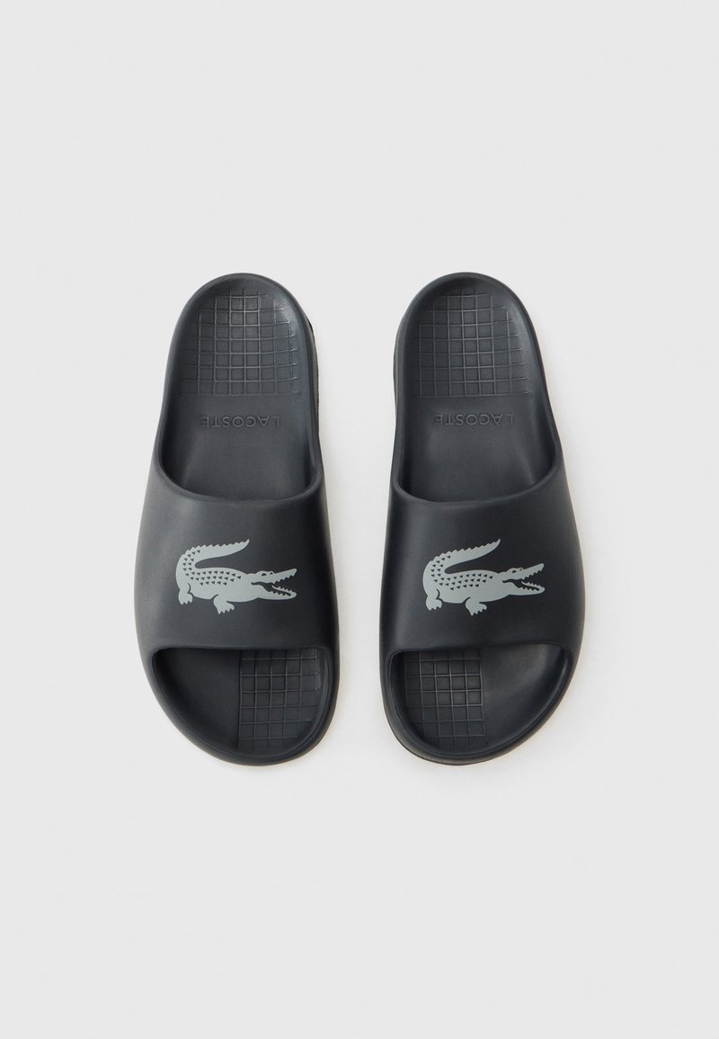 Des sandales noires en caoutchouc avec un design à bout ouvert, présentant une semelle texturée et un accent logo crocodile gris sur la lanière supérieure.