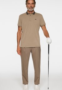 Mężczyzna ubrany w beżową koszulkę polo, pasujące spodnie, białe trampki i rękawiczkę golfową, trzymający kij golfowy na białym tle.