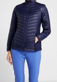 Marinblå vadderad jacka med dragkedja framtill, utrustad med vadderade paneler, ställbar krage och långa ärmar. Kombineras med blå byxor.