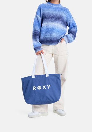 Personne portant un pull bleu dégradé et un pantalon beige, tenant un grand sac fourre-tout bleu avec un logo blanc "ROXY" et des poignées blanches.