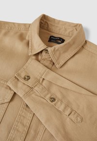 Beige Hemd mit Kragen, aus strukturiertem Stoff, mit zwei Brusttaschen und braunen Knöpfen. Etikett "CALLIOPE" sichtbar.