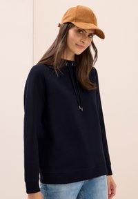 Marinblå sweatshirt med huva, dragsko och guldspetsade ändar. Matchad med en tanfärgad mockahatt. Mjuk textur, avslappnad design.