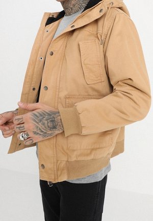 Hombre con chaqueta con capucha color beige con detalles de botones y cremallera, camisa gris y pantalones negros, mostrando manos y cuello tatuados.