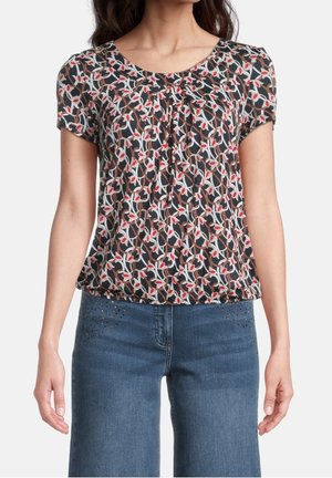 Kvinde iført sort top med korte ærmer med hvide og røde bladmønstre, kombineret med blå denimjeans med små pynt nær lommerne.