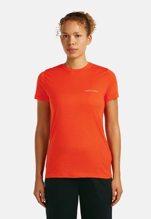Femme aux cheveux bouclés attachés en queue de cheval, portant un t-shirt à manches courtes orange vif uni avec le texte "icebreaker" et un pantalon noir, face à l'avant.
