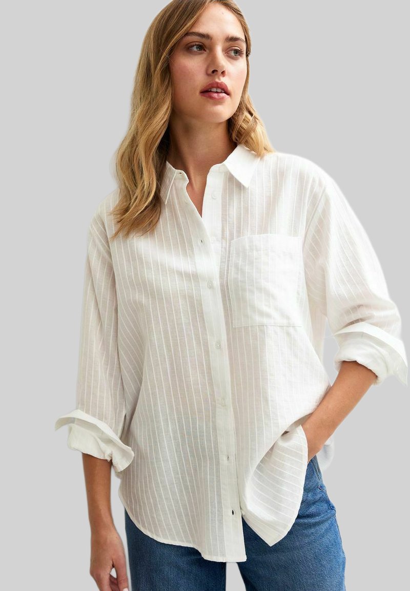 Camisa de botones de rayas blancas hecha de tejido ligero, con un bolsillo en el pecho, mangas enrolladas y un corte relajado. Combinada con vaqueros.