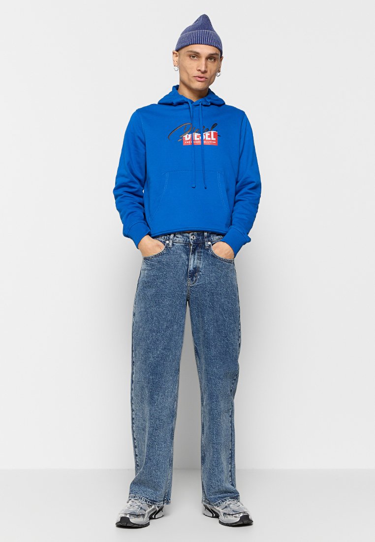 Sudadera azul con un logo en el frente, combinada con vaqueros de corte holgado en un tono azul claro y zapatillas grises. El modelo lleva un gorro azul.