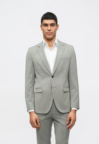 Ανθρακί κοστούμι με προσαρμοσμένη εφαρμογή, διαθέτοντας άνοιγμα με όχιτς, ενιαίο κλείσιμο με κουμπί και δύο μπροστινές τσέπες. Λευκό πουκάμισο από κάτω.