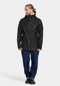 Svart vattentät jacka med hög krage, långa ärmar och dragkedja. I kombination med blå denimjeans och svarta skor.