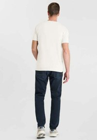 Uomo che indossa una maglietta bianca a maniche corte, jeans blu scuro e sneakers bianche, in piedi di spalle contro uno sfondo chiaro e semplice.