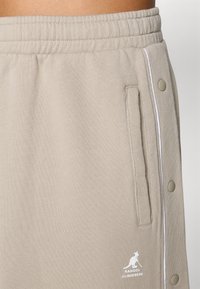 Beige jogger pants made of soft fabric, featuring side pockets, white trim, and snap button accents on the sides for adjustable fit.