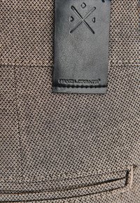 Teksturowana tkanina z brązowym i kremowym diagonalnym wzorem w jodełkę, z czarną skórzaną naszywką i tłoczonym logo na górze.
