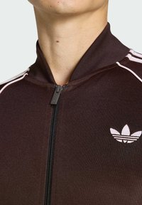 Mörkbrun zip-up jacka i slätt tyg, med vita ränder längs axlarna och en liten vit logotyp på bröstet.