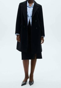 Manteau noir croisé à double boutonnage avec revers à encoches, associé à une chemise bleu clair boutonnée et une jupe noire. Escarpins noirs pointus avec des accents argentés.