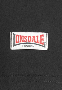 Fekete szövet, amelyen egy szőtt címke található, piros "LONSDALE" felirattal és fekete "LONDON" felirattal, fehér keretezéssel. Egyszerű, téglalap alakú dizájn.