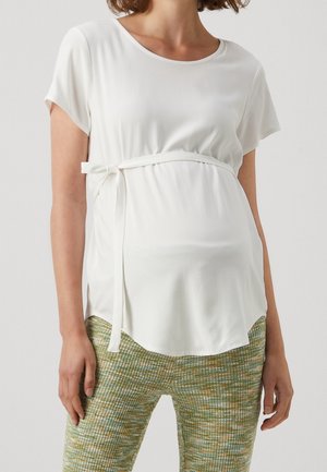 Woman wearing a white short-sleeve maternity top with side tie and green patterned pants, standing against a plain background.