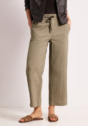 Kvinde iført beige teksturerede bukser med snøre, sort top, mørkebrun jakke og brune flade sandaler med flettede stropper.