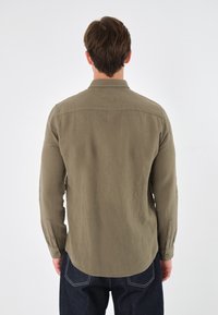 Man vänd bort, iklädd en olivfärgad långärmad skjorta med knappar och mörkblå jeans mot en vit bakgrund.