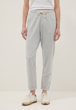 Persona que lleva pantalones deportivos gris claro con cordón, zapatillas blancas y una camiseta blanca corta, de pie contra un fondo claro y sencillo.