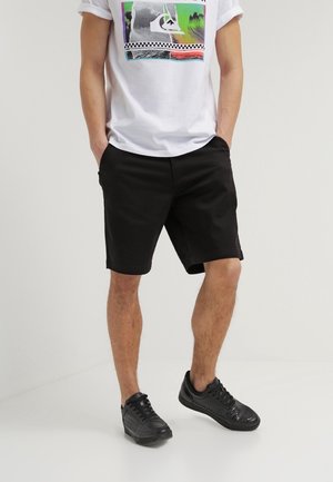 Mand iført en hvid t-shirt med tryk, sorte shorts og sorte sneakers står med hænderne i lommerne mod en ensfarvet baggrund.