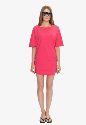 Kobieta w oversize'owych czarnych okularach przeciwsłonecznych, jasnoróżowej mini sukience z krótkim rękawem i czarnych klapkach, stojąca na białym tle.