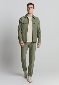 Olivgrüne Jacke mit zwei Brusttaschen, kombiniert mit passenden Hosen. Das Outfit wird mit einem cremefarbenen T-Shirt und beigen Sneakers abgerundet.