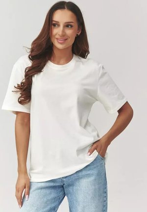 Kobieta o długich brązowych włosach, ubrana w prostą białą koszulkę i jasnoniebieskie jeansy, pozująca z jedną ręką w kieszeni na jednolitym tle.