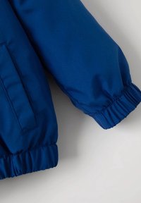 Modrá nylonová bunda s elastickým manžetovým rukávem a boční kapsou. Hladká textura, mírně lesklý povrch. Jednoduchý design bez viditelného kování.