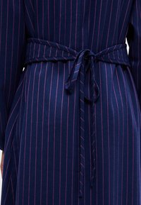 Kék csíkos ruha rózsaszín kiegészítőkkel, megkötős derékszíjjal, hosszú ujjal és puha textúrájú anyagból készült. A függőleges csíkok vizuális érdekességet adnak.