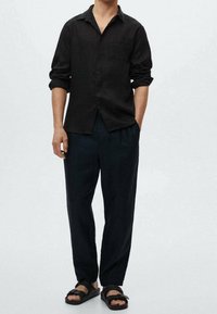 Camisa de lino negra con mangas enrolladas, abotonada, combinada con pantalones oscuros de corte holgado y sandalias negras de diseño con tira.