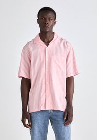 Kortermet rosa stripete skjorte i lett stoff med lomme. Har klassisk krage og hvite knapper, båret med blå jeans.
