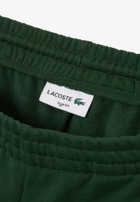 Pantalons de survêtement vert ajustés avec une taille côtelée. Une étiquette présentant le logo Lacoste est visible, indiquant les détails de la marque et de l'ajustement.