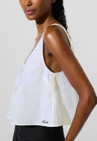 Ženska, oblečena v brezrokavnik, bel kratek top z ohlapnim krojem in majhnim vezenim logotipom "Karl" blizu roba, kombiniran s črnimi hlačami.