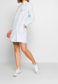 Light blue denim shirt dress with long sleeves, button-down front, two chest pockets, and side pockets, paired with white sneakers.