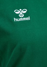 Camiseta de algodón verde con un logo bordado en blanco que presenta un diseño de abeja y la palabra "hummel" debajo, mostrando una textura suave.