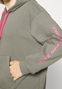 Un sweat à capuche vert olive avec des cordons de tirage rose, une poche avant et un logo Nike rose sur la manche. Fait en tissu doux avec une coupe décontractée.
