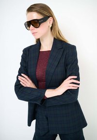 Blazer a quadri neri con un design strutturato, abbinato a un top color vinaccia. Occhiali da sole marroni oversize e orecchini con dettagli dorati completano il look.