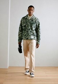 Uomo che cammina all'interno indossando una giacca in pile verde a motivo floreale, pantaloni beige, sneakers nere e bianche, mentre tiene una borsa per fotocamera nera.