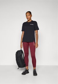 Sort t-shirt med "TERREX" logo, bordeaux leggings og sort rygsæk. Modellen har sorte træningssko med tekstureret overflade. Enkel design.