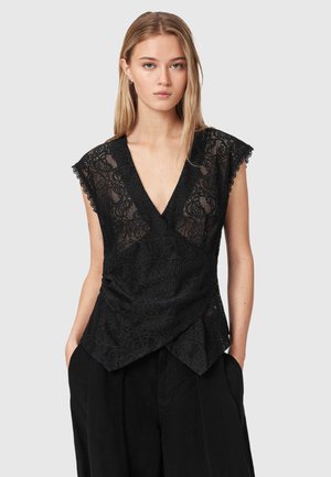 Jeune femme aux longs cheveux blonds portant un top noir en dentelle sans manches avec un col en V et un pantalon noir, debout, les mains dans les poches.